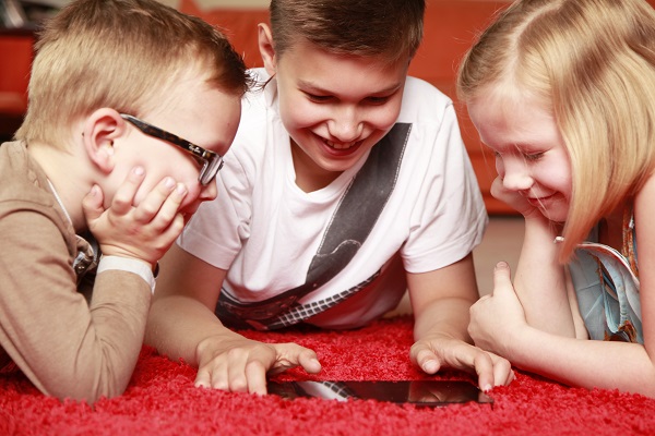 Projekt "Spieleratgeber-NRW". Drei Kinder liegen auf einem Teppich um ein Smartphone herum und betrachten es.