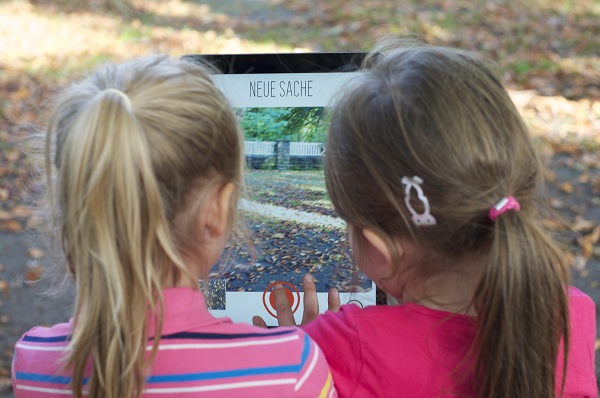 Projekt "#stadtsache - crossemdiale Teilhabe an der Stadt". Zwei Mädchen betrachten auf einem Tablet ein Bild eines Weges.