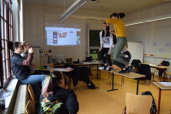 Foto eines Klassenraums. Auf der Heizung am Fenster sitzen zwei Jugendliche. Einer von ihnen hält ein Smartphone in der Hand, welches auf drei weitere Jugendliche gerichtet ist, die von einem Tisch springen.