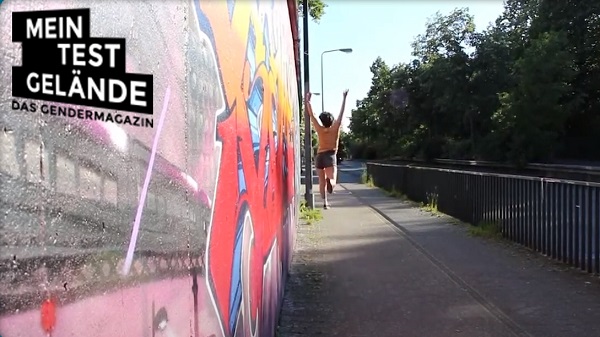 Foto des Projekts "meinTestgelände - das Gendermagazin". Auf einem Gehweg neben einer mit Graffiti besprühten Mauer springt eine Person mit nach oben gestreckten Armen.