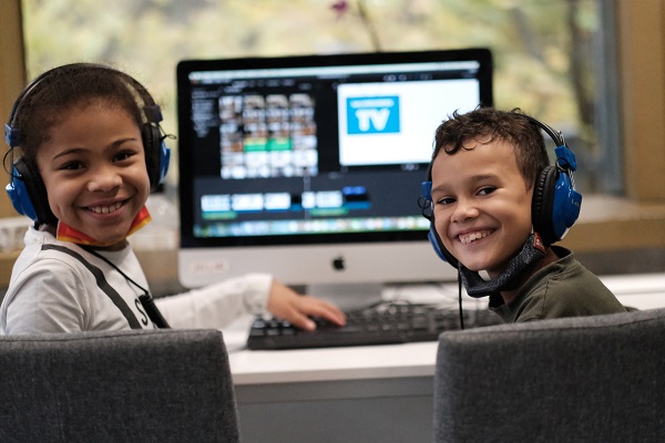 Foto vom Projekt "Salzbrunner TV": zwei Kinder sitzen vor einem Computerbildschirm, auf welchem ein Schnittprogramm geöffnet ist, tragen Kopfhörer und drehen sich mit ihrem Gesicht zur Kamera und lächeln.