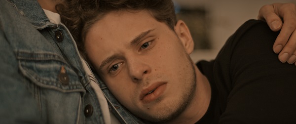 Screenshot aus einer Folge der Serie "Kuntergrau". Großaufnahme des Gesichts eines jungen Mannes, der nachdenklich schaut und in den Armen einer anderen Person liegt.