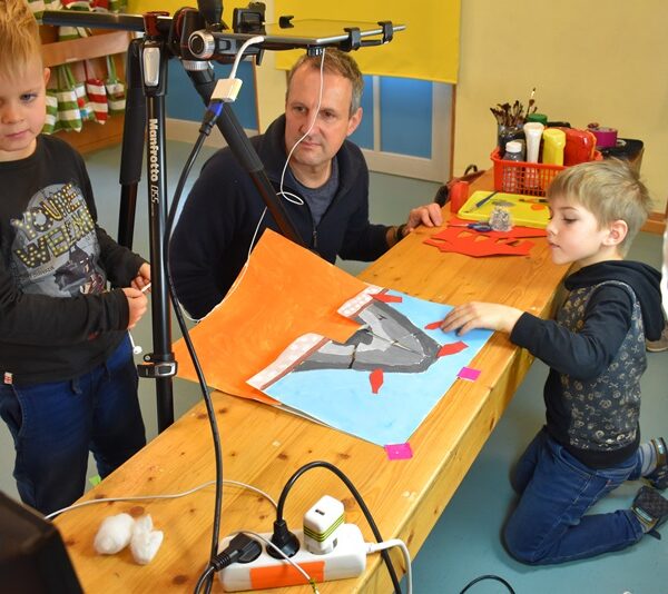 Zwei Kita-Kinder und ein Erzieher fotografieren ein selbstgemaltes Bild