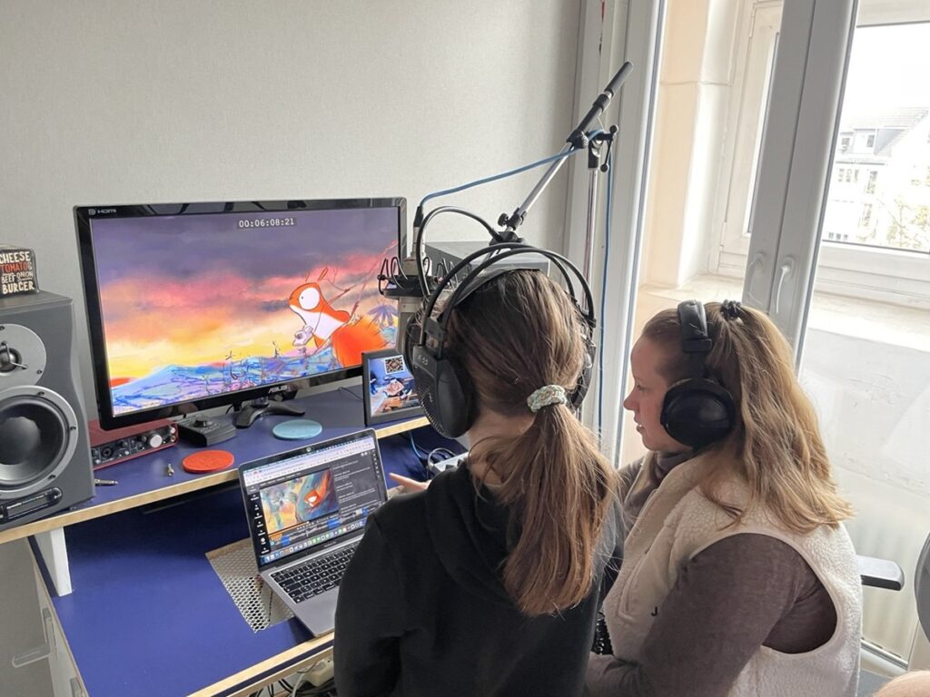 Zwei Jugendliche sitzen nmit Kopfhörern vor einem Laptop und einem Bildschirm